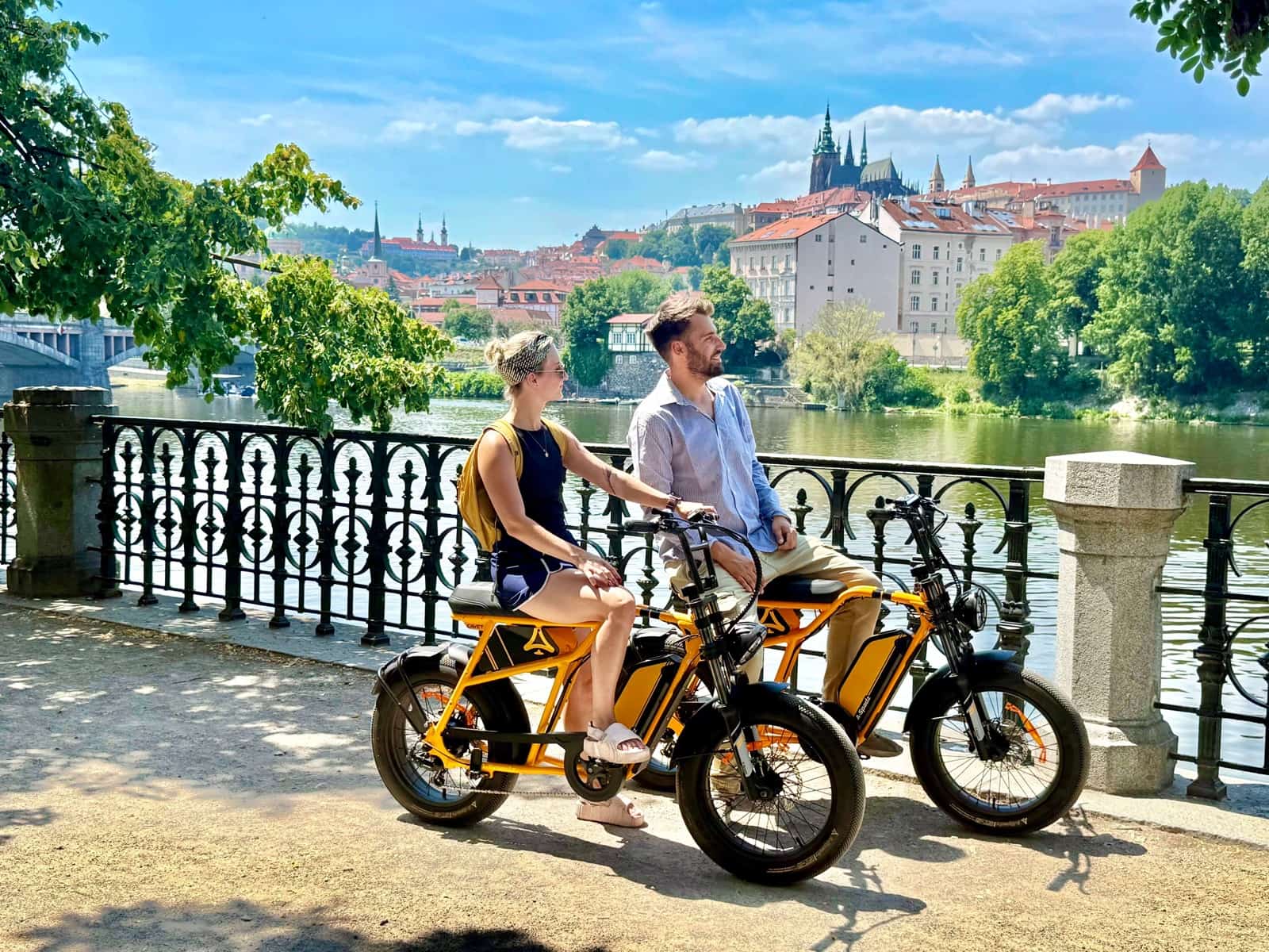 Dvojice na oranžových chopper e-kolech u Vltavy s Pražským hradem v pozadí