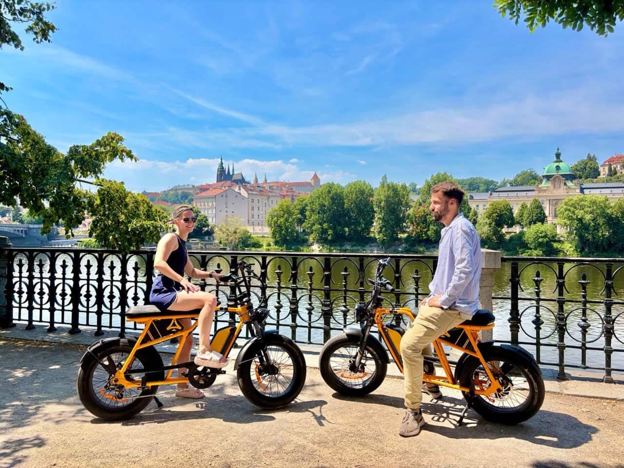 Twee rijders op oranje chopper e-bikes aan de rivier met de Praagse Burcht erachter