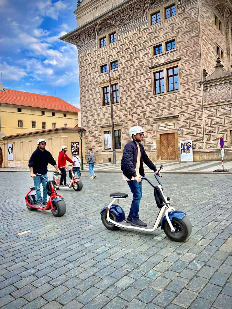 Riders standing on SCROOSER e-scooters over cobblestones by the Schwarzenberg Palace
